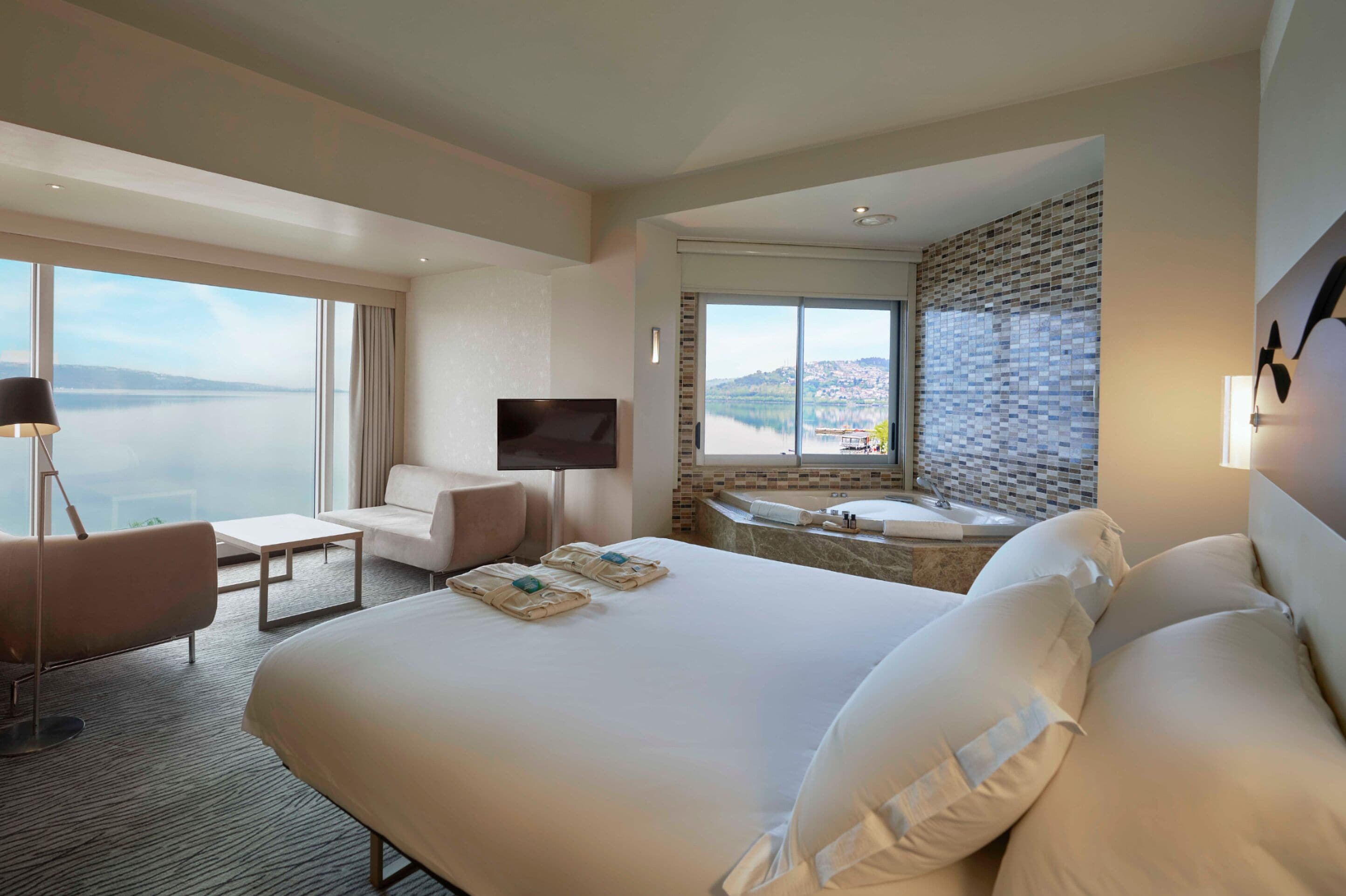 Quarto de hotel com uma cama grande, área de estar com sofá e poltrona, televisão de tela plana, banheira e vista para a cidade e para a água.