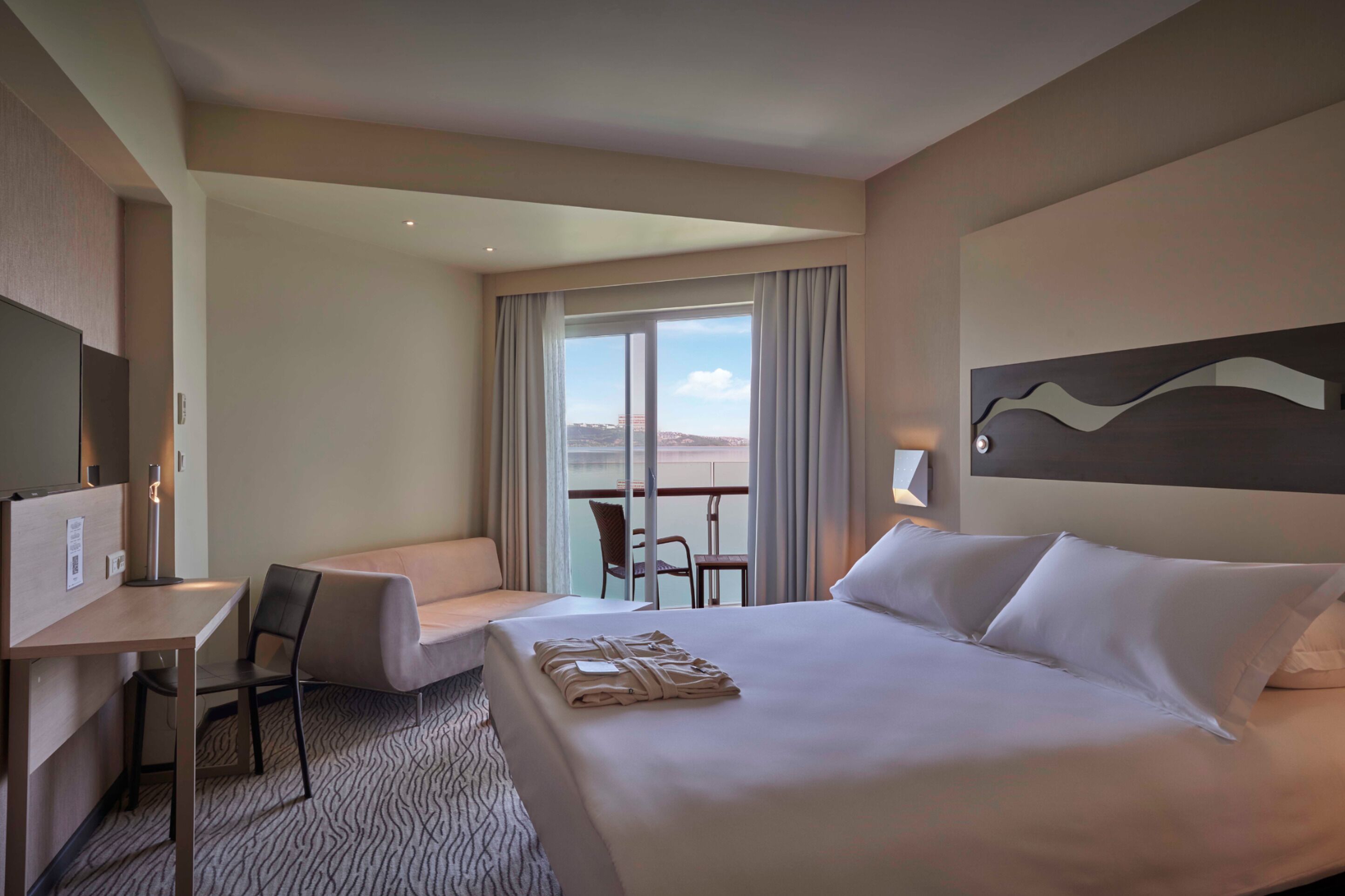 A hotel room with a large bed, a desk, a chair, and a balcony with a view.