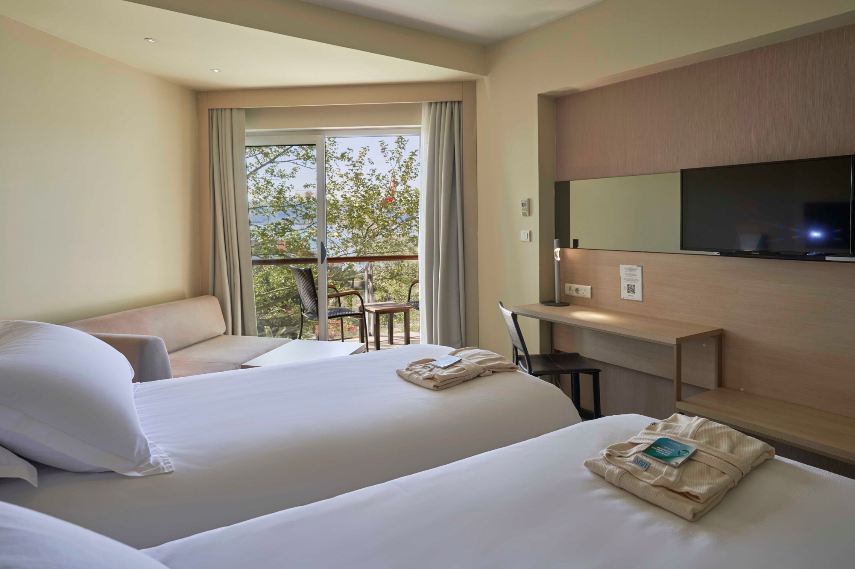 A hotel room with two beds, a desk, a chair, a TV, and a balcony with a view of trees.