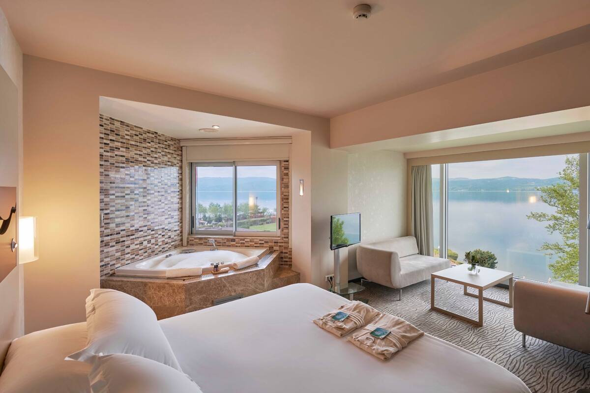 A hotel room with a large bed, a bathtub, a sofa, a coffee table, and a TV. The room has a view of a lake and mountains.