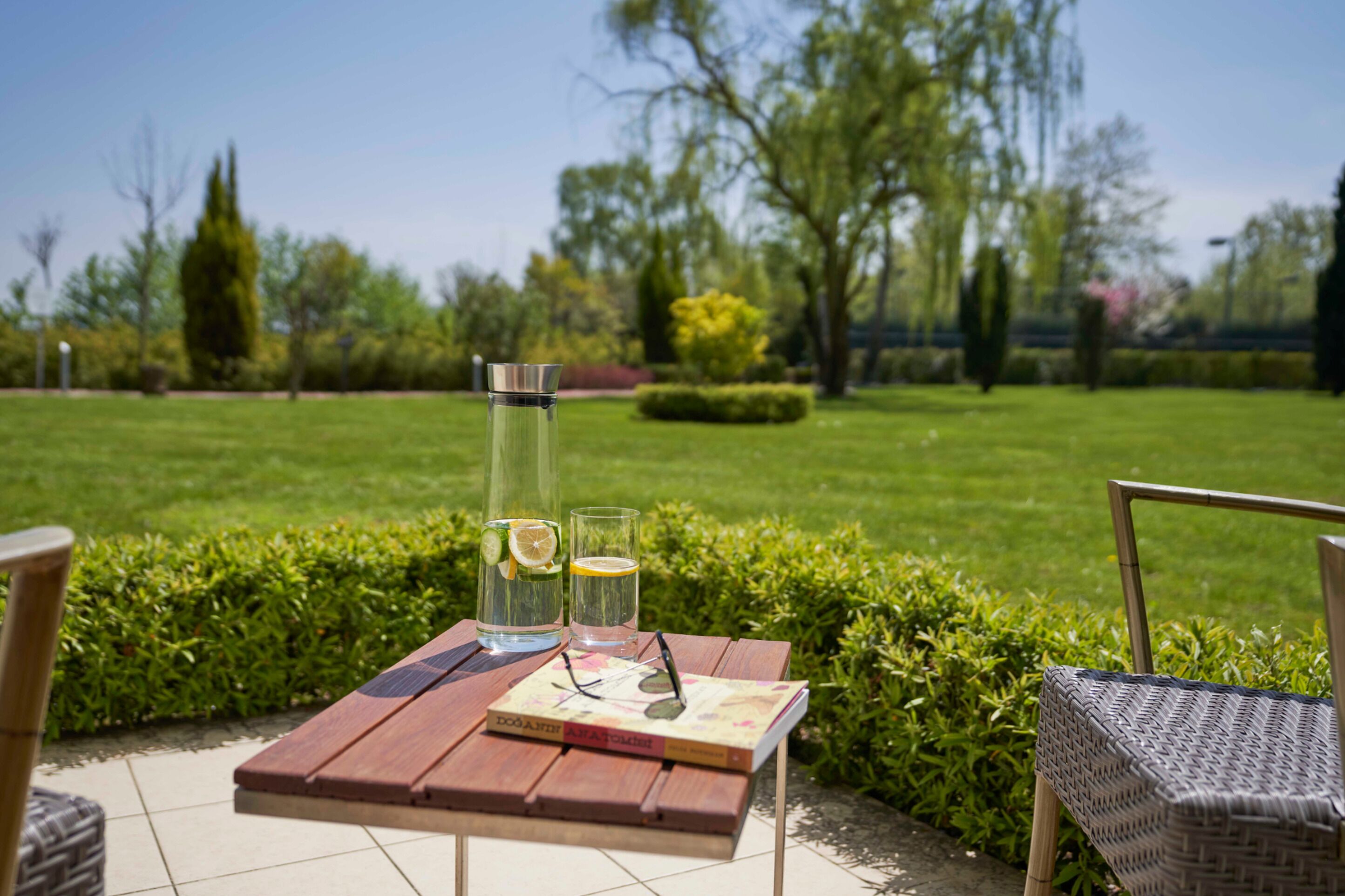 Una mesa de madera con un libro, un vaso y una botella en un jardín.