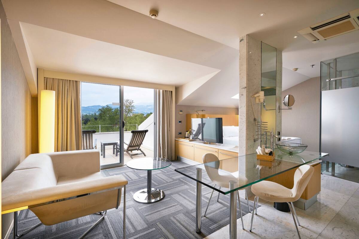 A modern hotel room with a glass dining table, a sofa, a TV, and a bathroom with a glass shower.