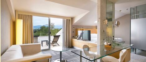 Habitación de hotel moderna con una mesa de comedor de vidrio, un sofá, un televisor y un baño con cabina de ducha de vidrio.