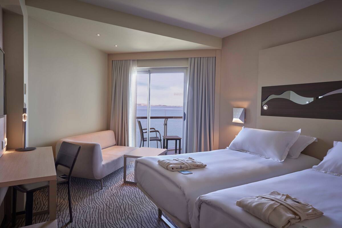 A hotel room with two beds, a desk, a chair, and a view of the sea through a large window.