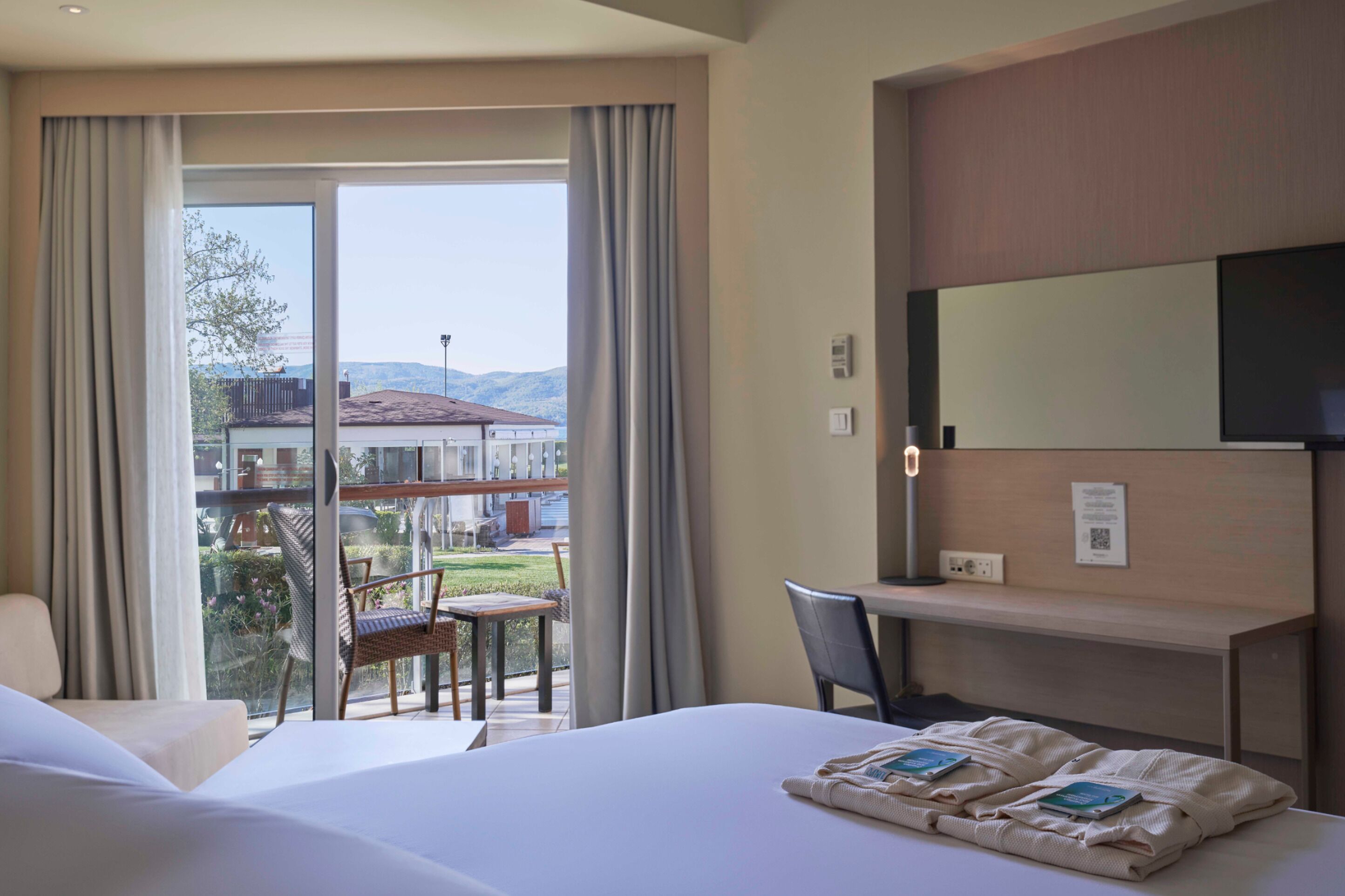 A hotel room with a bed, a desk, and a chair. There is a large window with curtains, and a view of the outside.