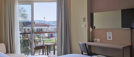 A hotel room with a bed, a desk, and a chair. There is a large window with curtains, and a view of the outside.