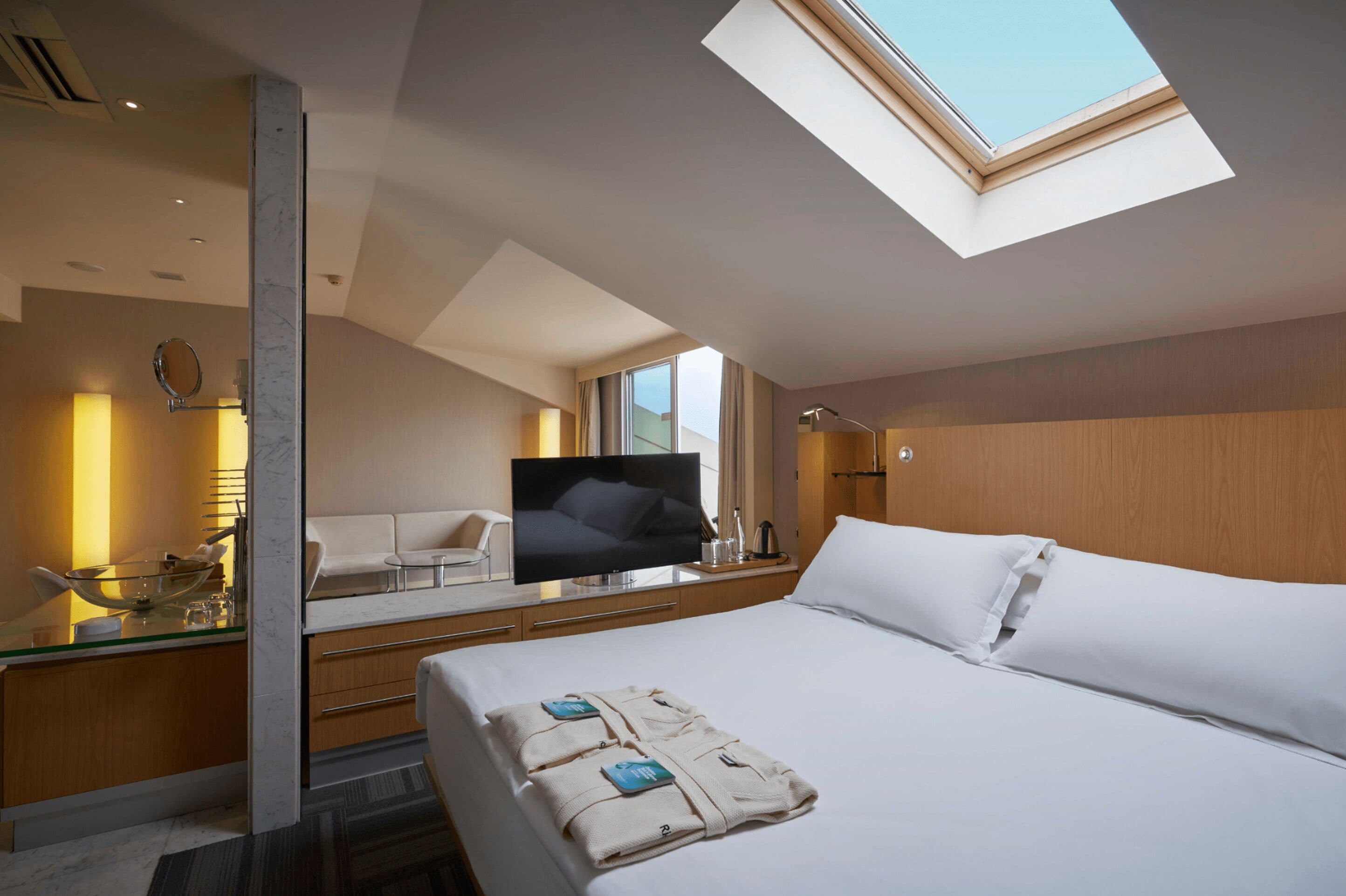 A modern hotel room with a large bed, a television, a glass-top desk with a bowl and glasses, and a skylight.