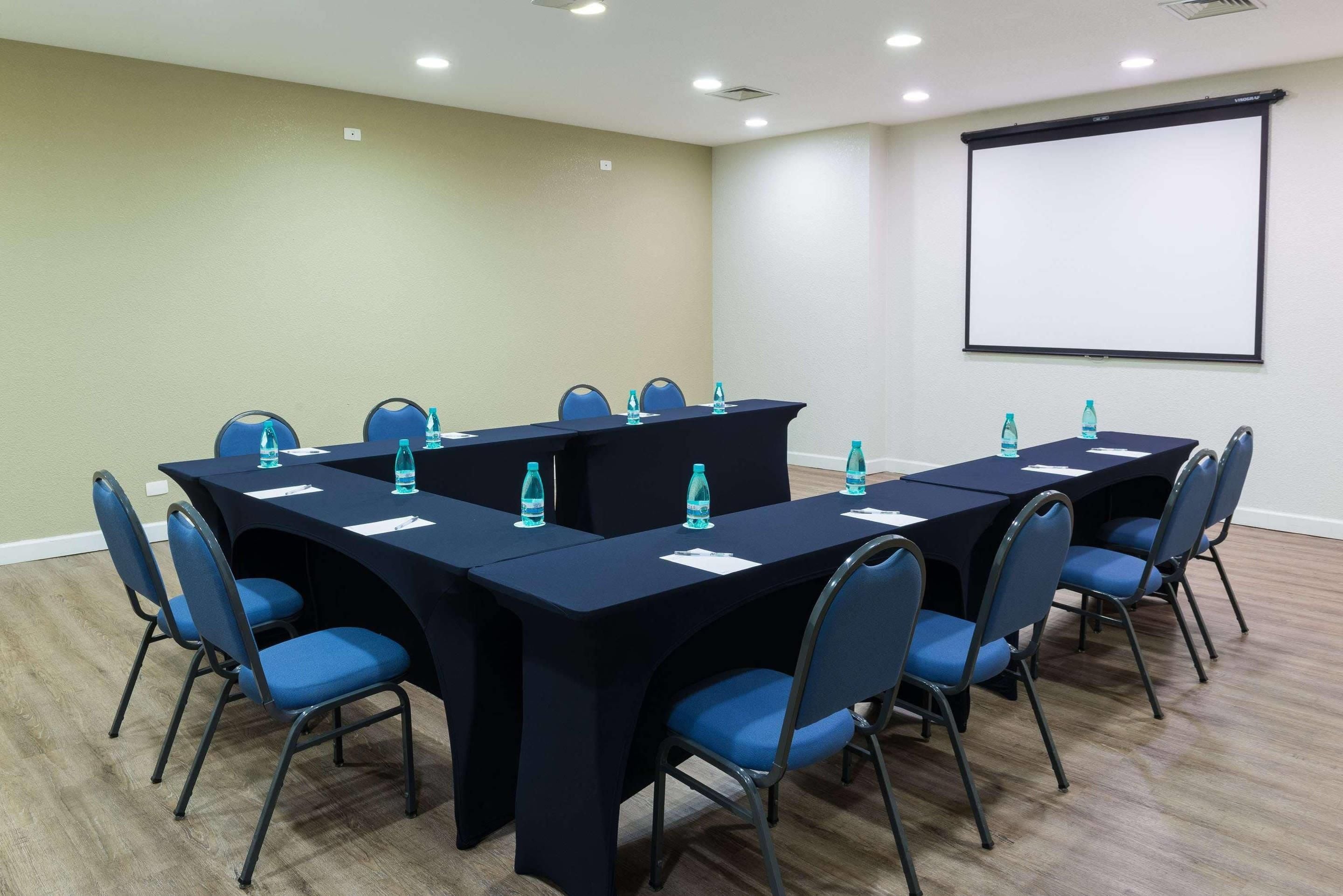 A conference room with a long table, chairs, water bottles, and a projector screen.