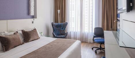 Habitación de hotel con una cama grande, un escritorio, una silla, un televisor y una ventana con cortinas.