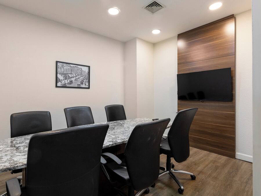Un salón de conferencias con una larga mesa, sillas negras, un cuadro enmarcado en la pared y un panel de madera a la derecha.