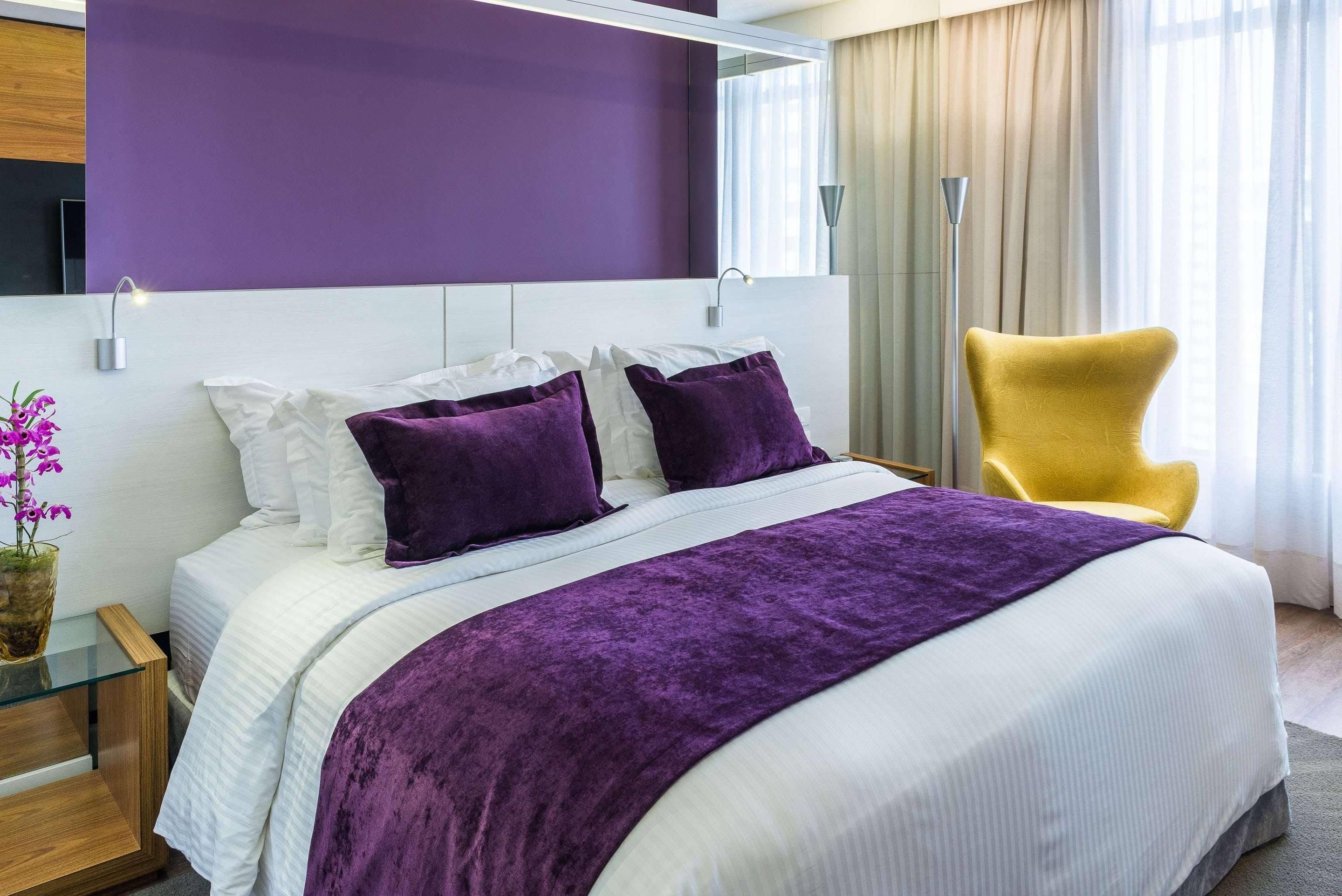 A modern hotel room with a large bed, purple bedding, a yellow armchair, and a view of the outdoors.