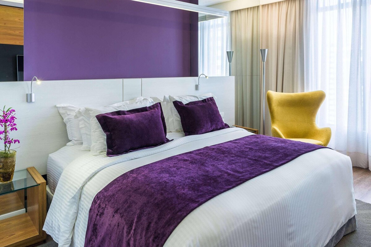 A modern hotel room with a large bed, purple bedding, a yellow armchair, and a view of the outdoors.