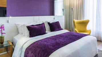 A modern hotel room with a large bed, purple bedding, a yellow armchair, and a view of the outdoors.