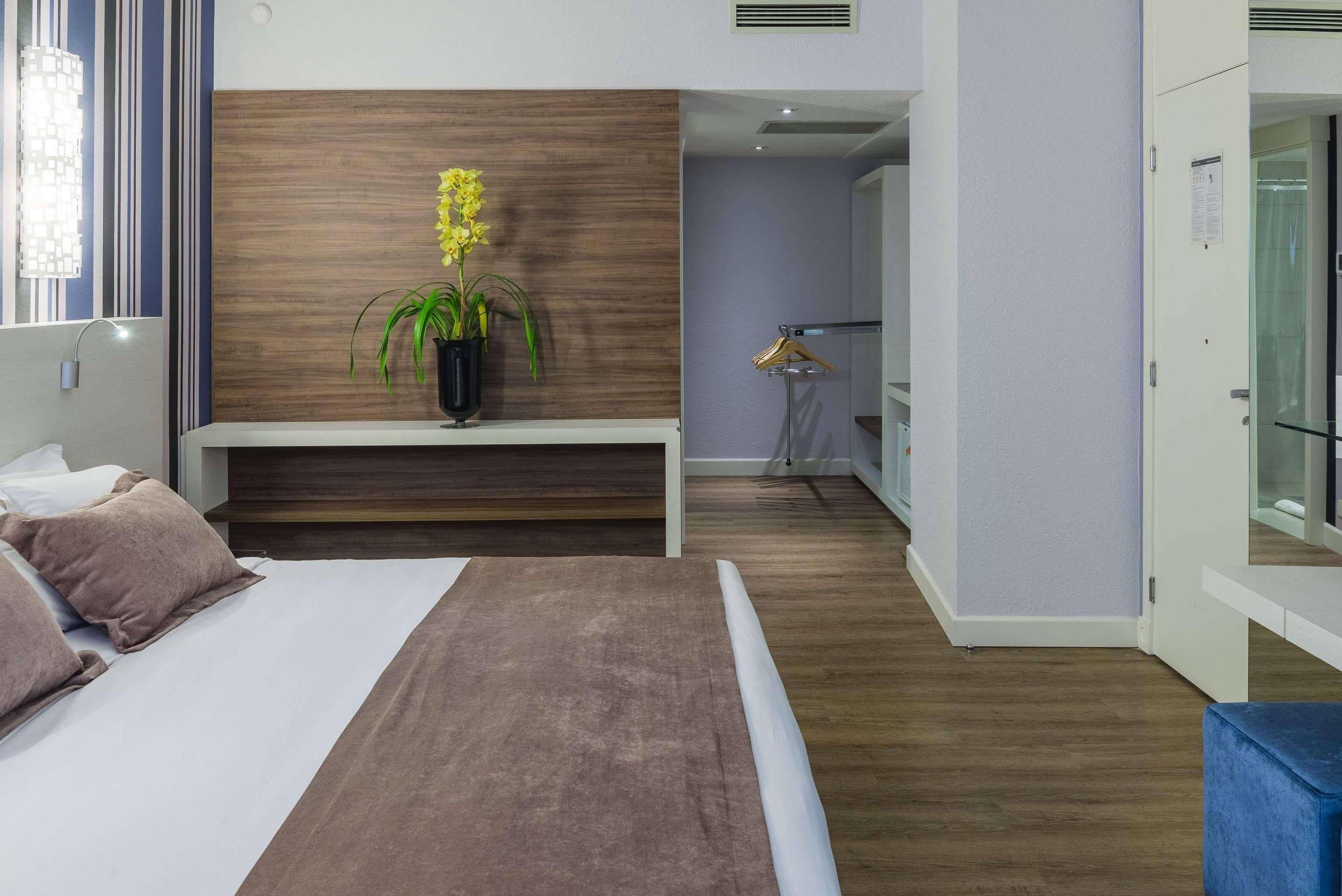 A modern hotel room with a bed, a bedside table, a potted plant, and a view of the hallway.