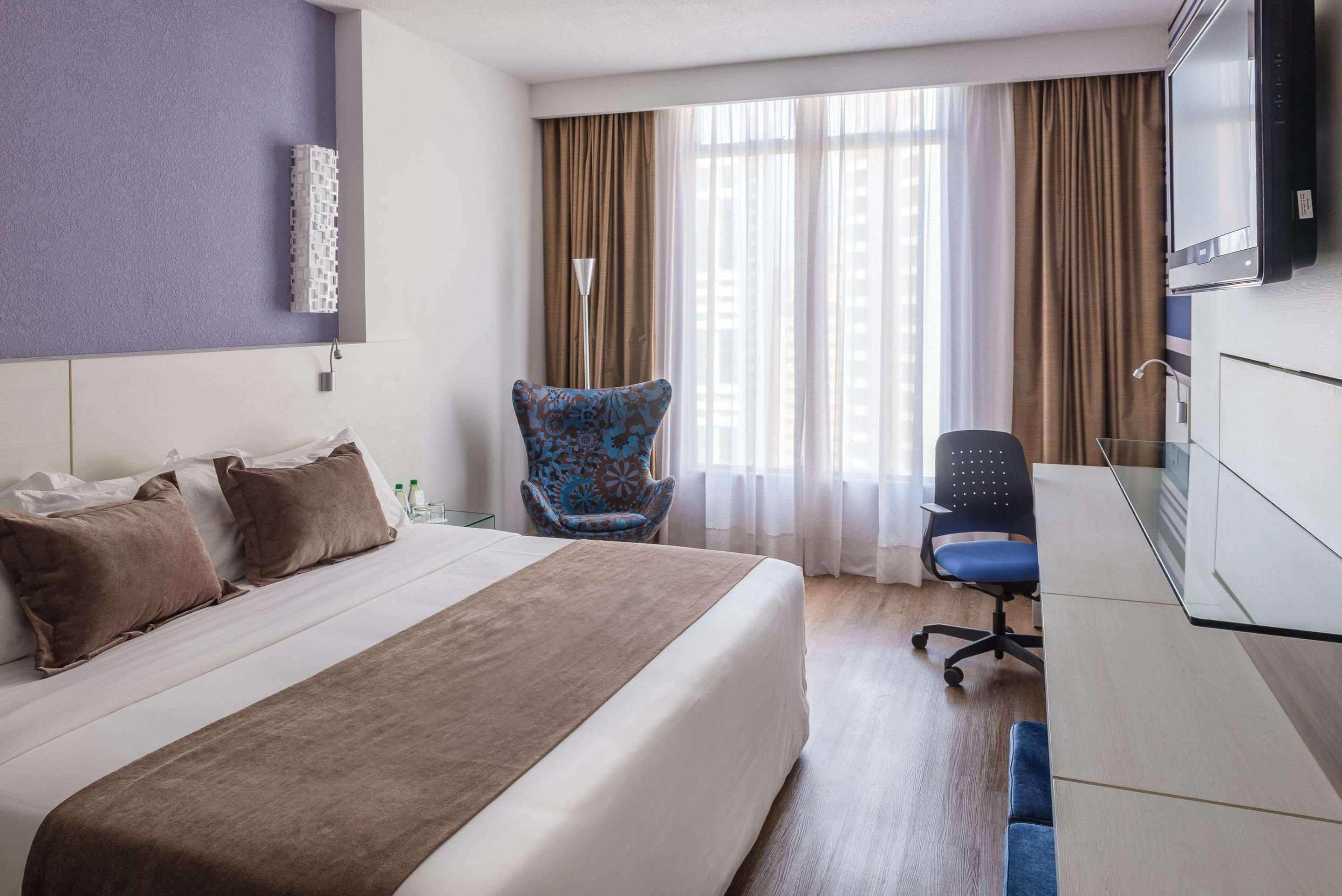 A hotel room with a bed, a desk, a chair, and a large window with curtains.