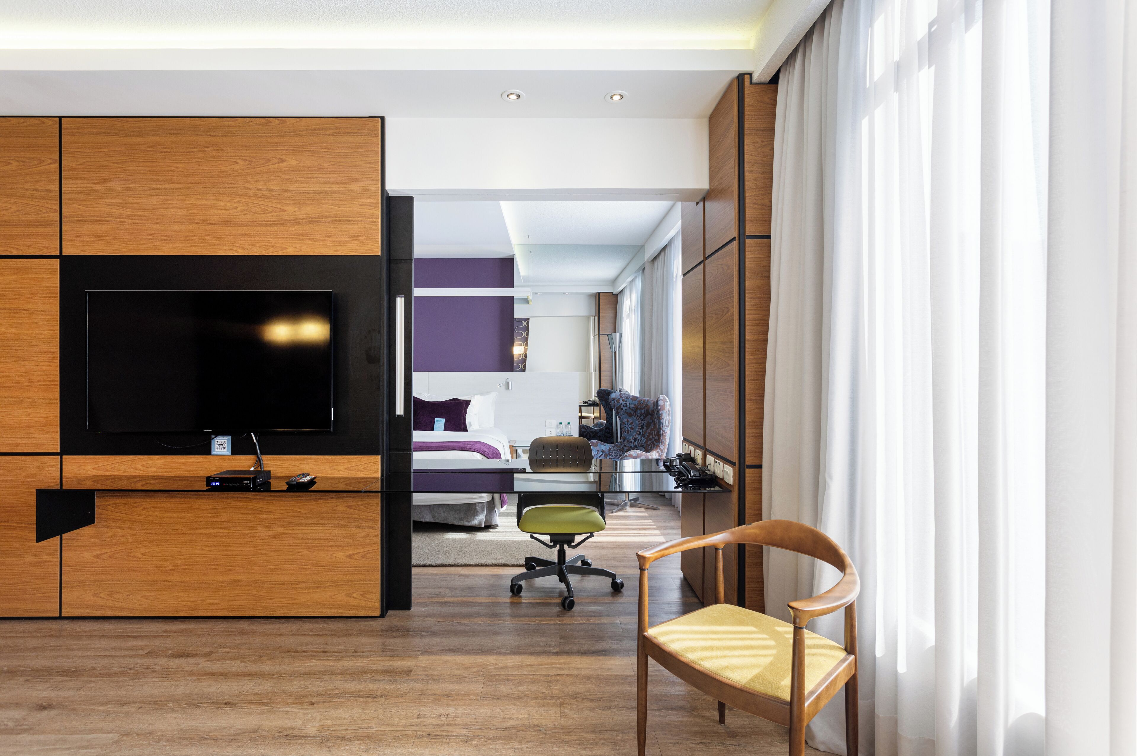 A modern hotel room with a large flat-screen TV mounted on a wooden panel, a desk with a computer, a chair, and a bed with purple bedding.
