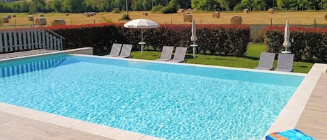 Una piscina con agua azul cristalina, rodeada por una terraza de madera. Hay sillones de descanso y sombrillas, y se ve un paisaje verde con colinas al fondo.