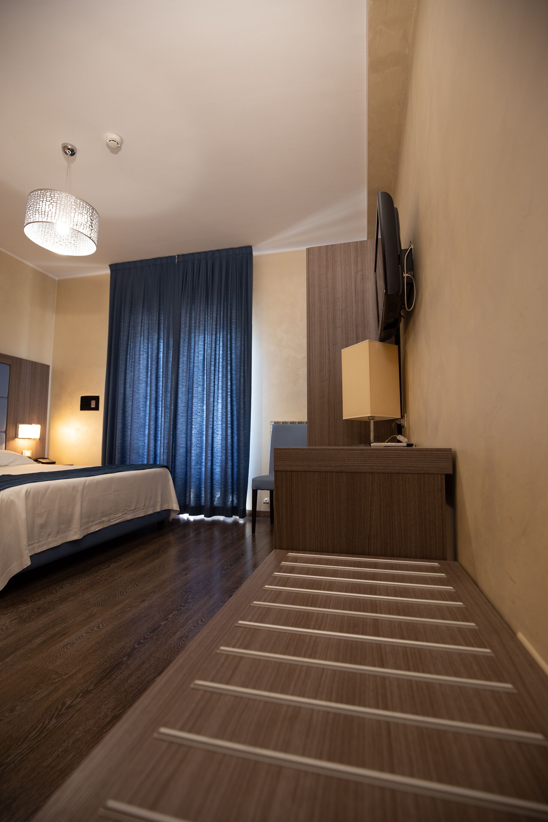 Una habitación de hotel con cama, escritorio, silla, televisión y un ventanal con cortinas.