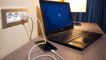 A laptop on a wooden desk with a power outlet and a lamp in the background.