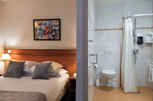 A hotel room with a bed, pillows, a nightstand, a lamp, and a framed abstract painting on the wall.