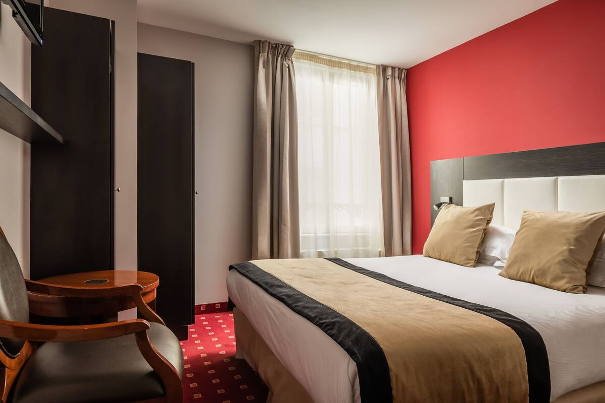 Ett hotellrum med en säng, en trästol, ett litet bord, en garderob och ett fönster med gardiner.