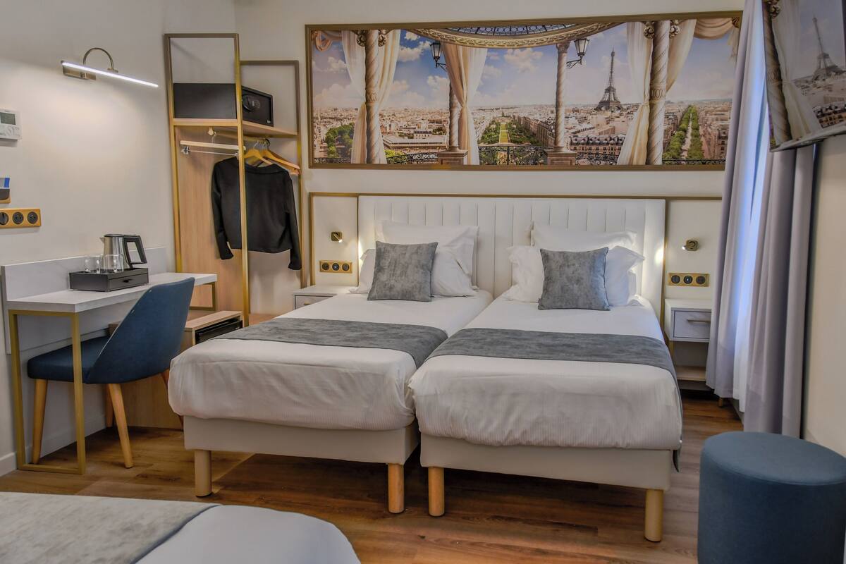 Ett hotellrum med två sängar, ett skrivbord, en stol, en garderob och en stor väggmålning som föreställer en stadsbild.