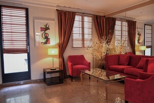 Uma sala de estar moderna com um sofá vermelho, uma mesa de centro de vidro e uma poltrona vermelha. Há grandes janelas com cortinas marrons, um abajur e um vaso decorativo com ramos.