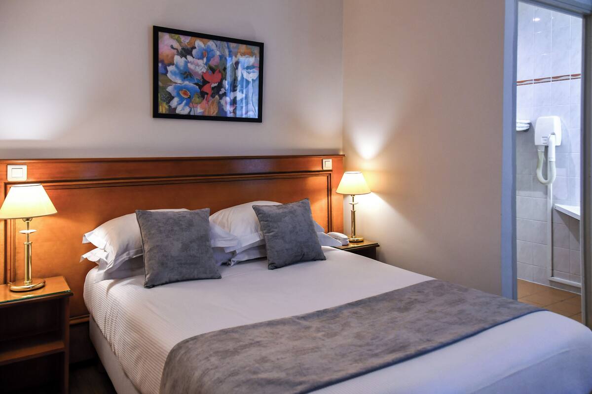 Ett hotellrum med en stor säng, två sänglampor, ett inramat konstverk med blommotiv på väggen och ett badrum som syns genom en öppen dörr.