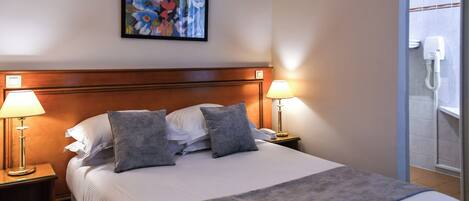 Habitación de hotel con una cama grande, dos lámparas de noche, una obra de arte floral enmarcada en la pared y un baño visible a través de una puerta abierta.