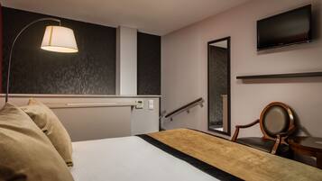 Una habitación de hotel con cama, lámpara de noche, televisor, sillón y espejo.