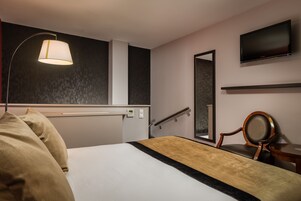 Una habitación de hotel con cama, lámpara de noche, televisor, sillón y espejo.