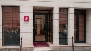 Entrada de un hotel con puerta de vidrio, un letrero rojo con una 'H' blanca y tres estrellas, y felpudo con el nombre del hotel.