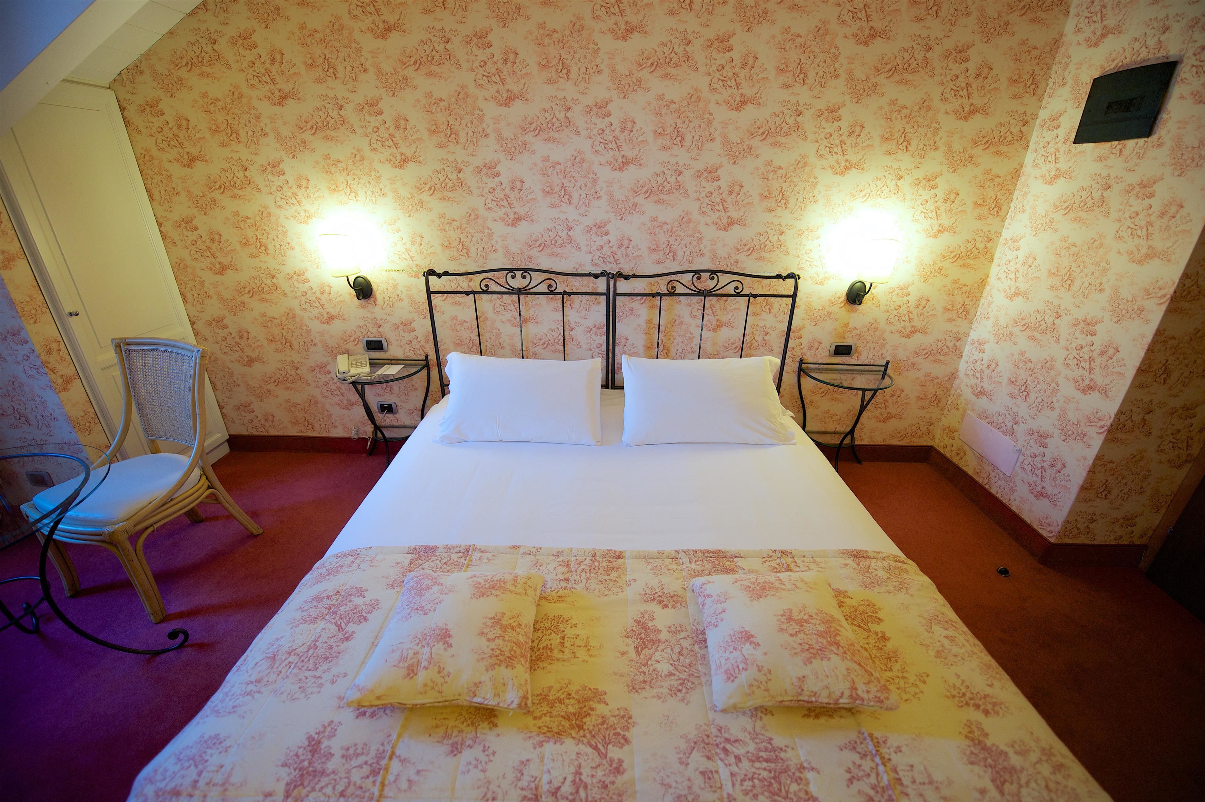 Cama bem arrumada com lençóis brancos, duas luminárias de cabeceira e um papel de parede estampado com flores.