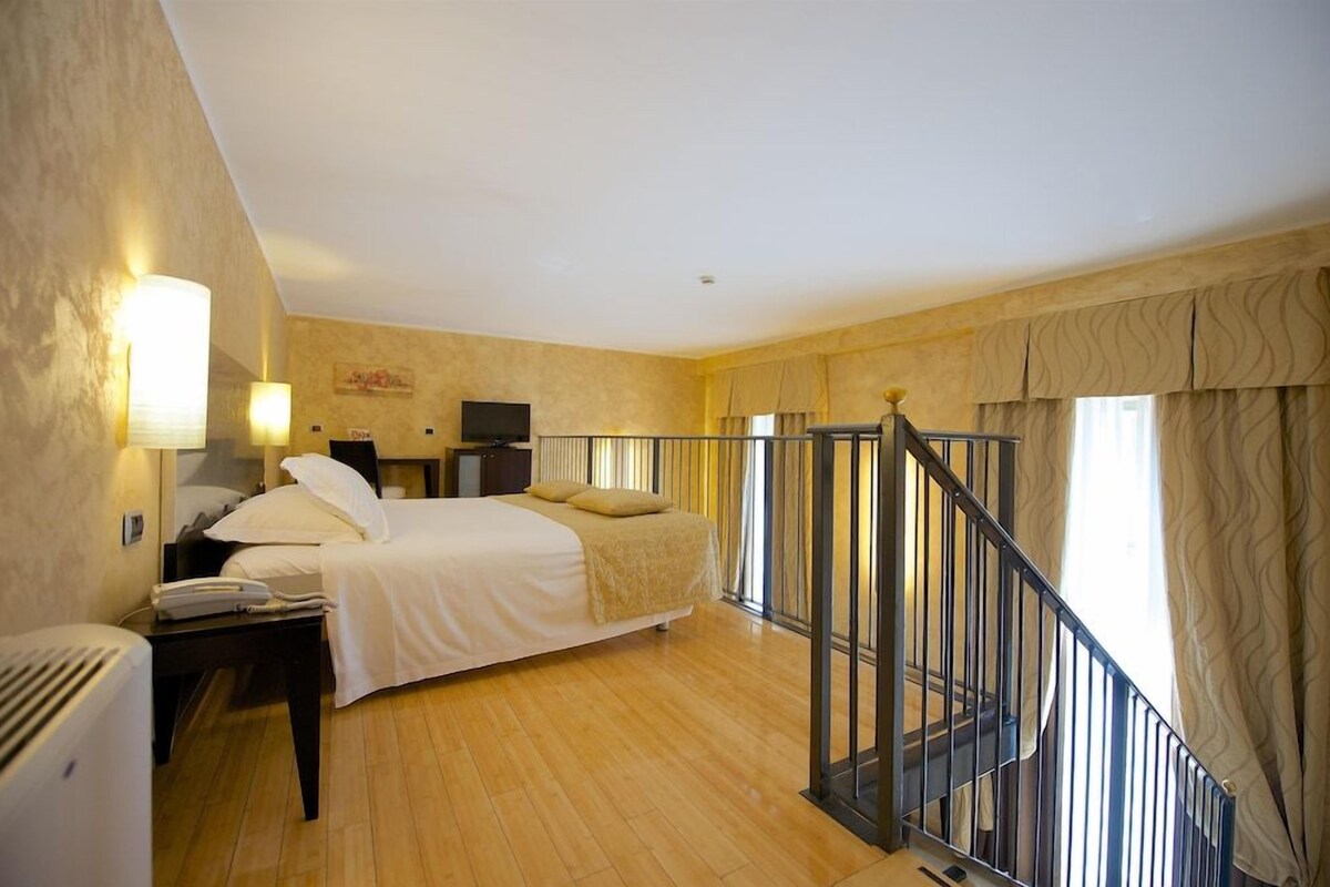 A hotel room with a bed, bedside table, lamp, and a small desk with a computer.