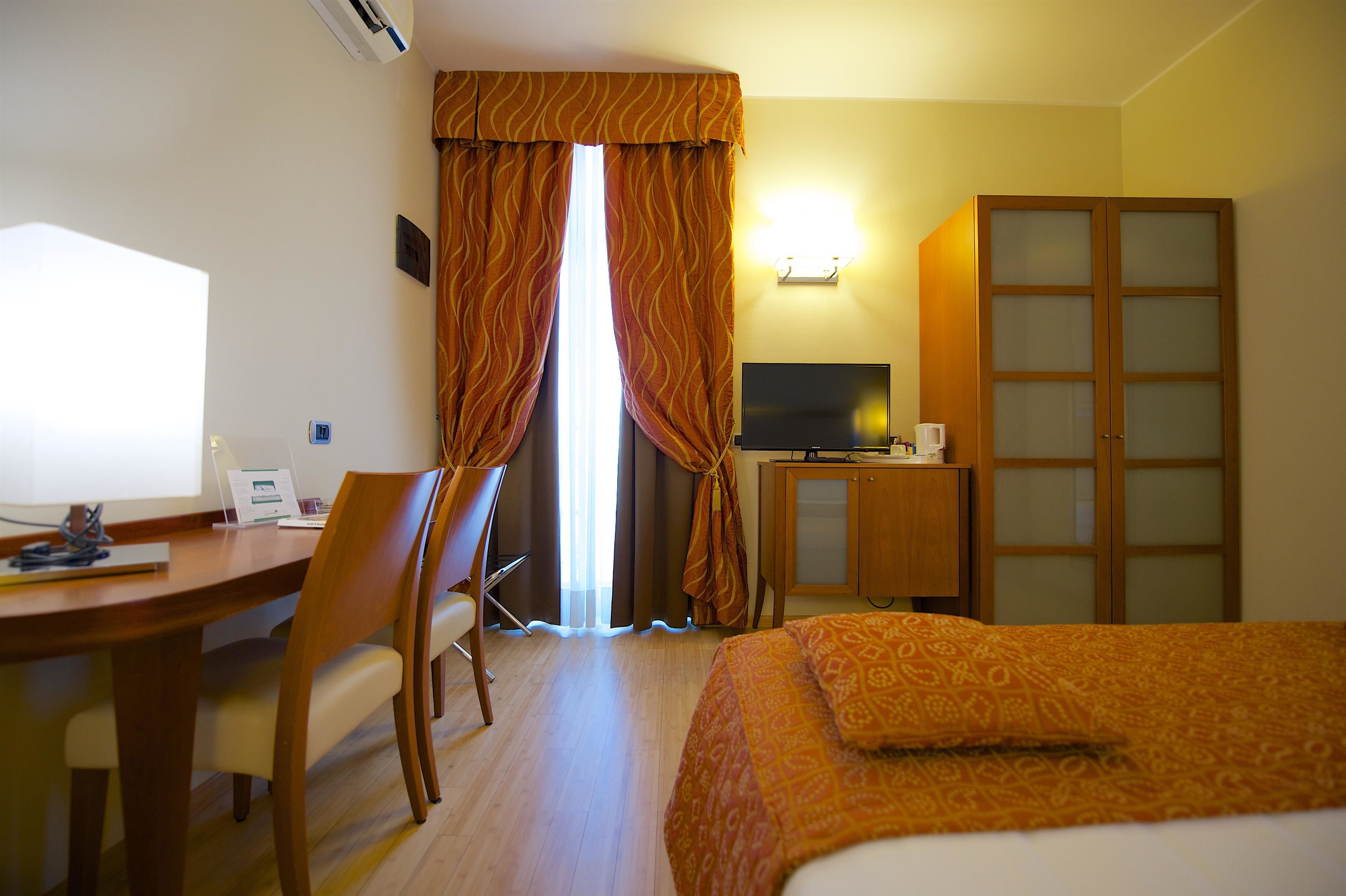 A hotel room with a bed, desk, chair, television, wardrobe, and curtains.