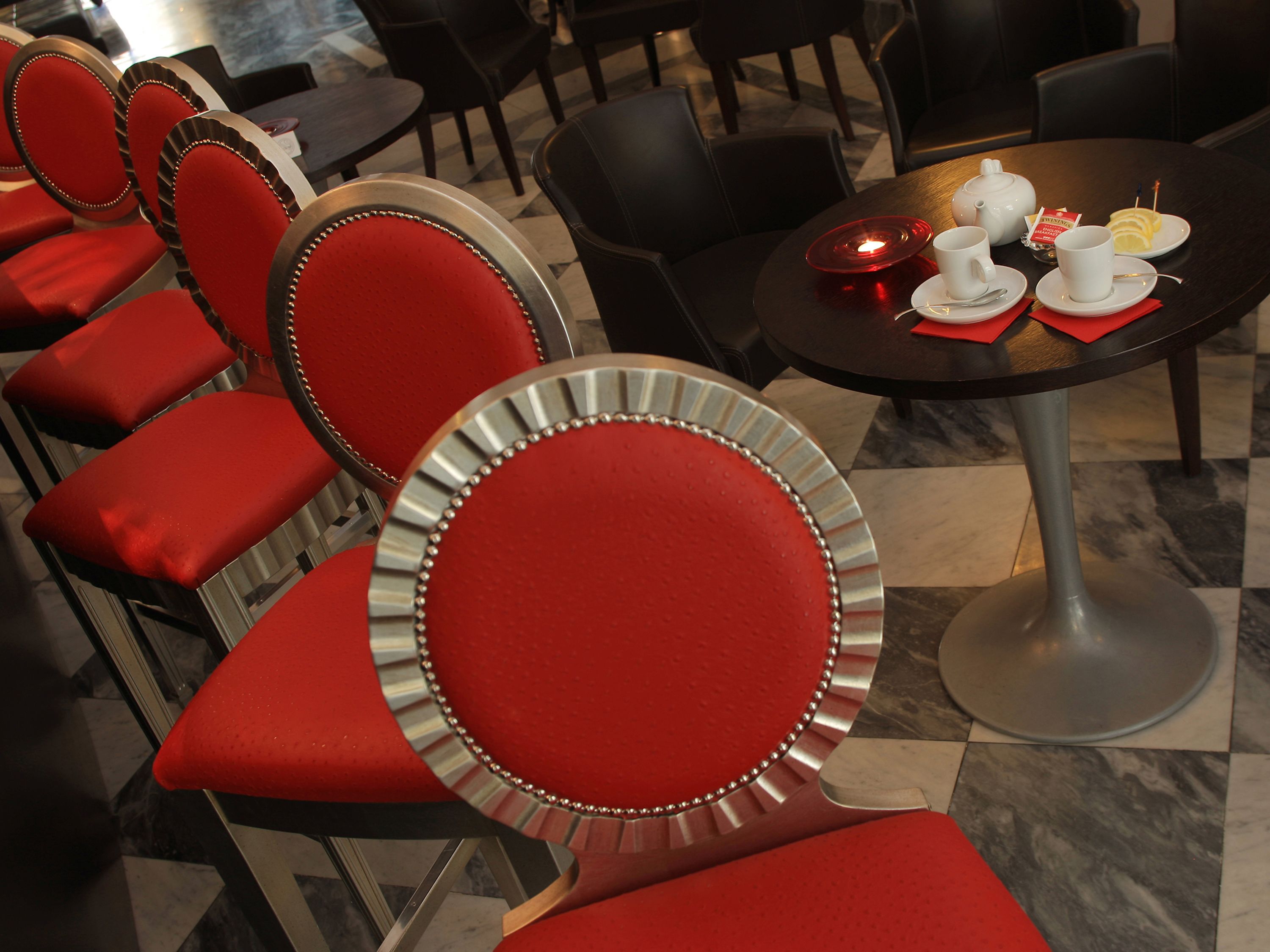 A restaurant interior with red upholstered chairs and a table set with white cups, a teapot, and a plate of sliced fruit.