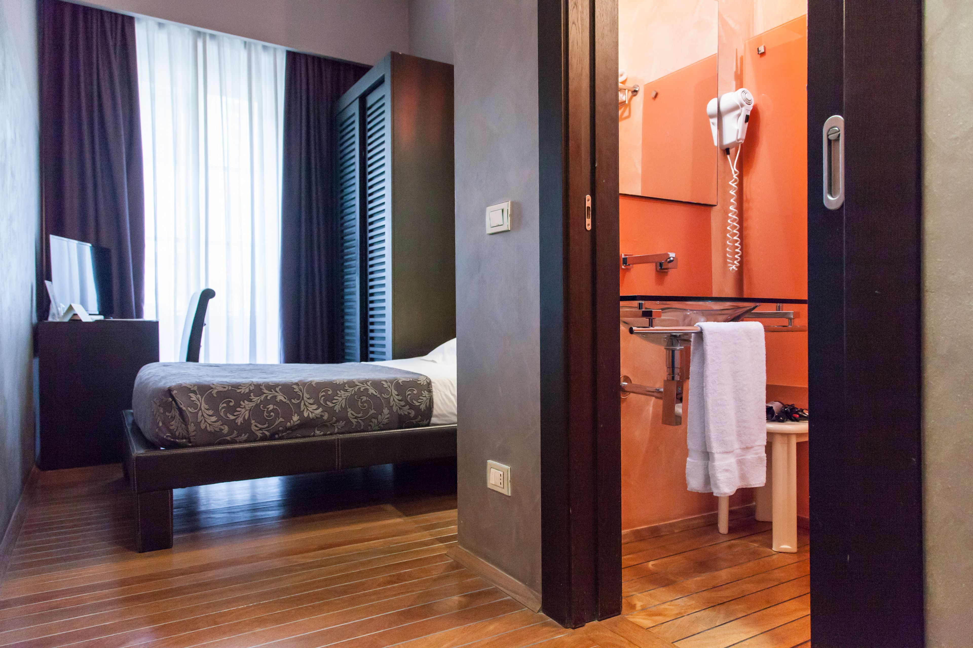 A hotel room with a bed, a desk, a chair, a window with curtains, and a bathroom with a towel rack and a hairdryer.