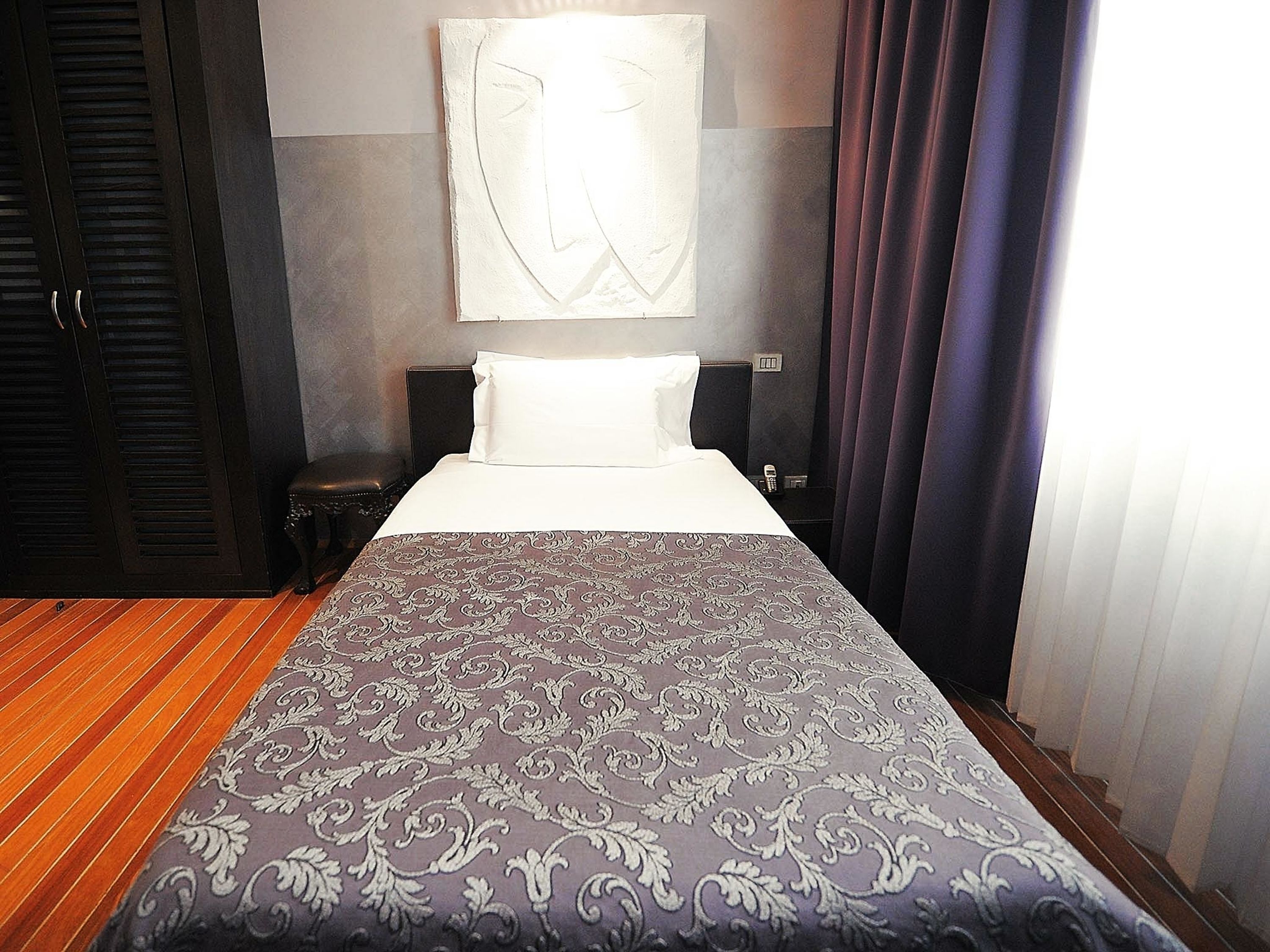 A hotel room with a large bed, a nightstand, a wall-mounted artwork, and a window with curtains.