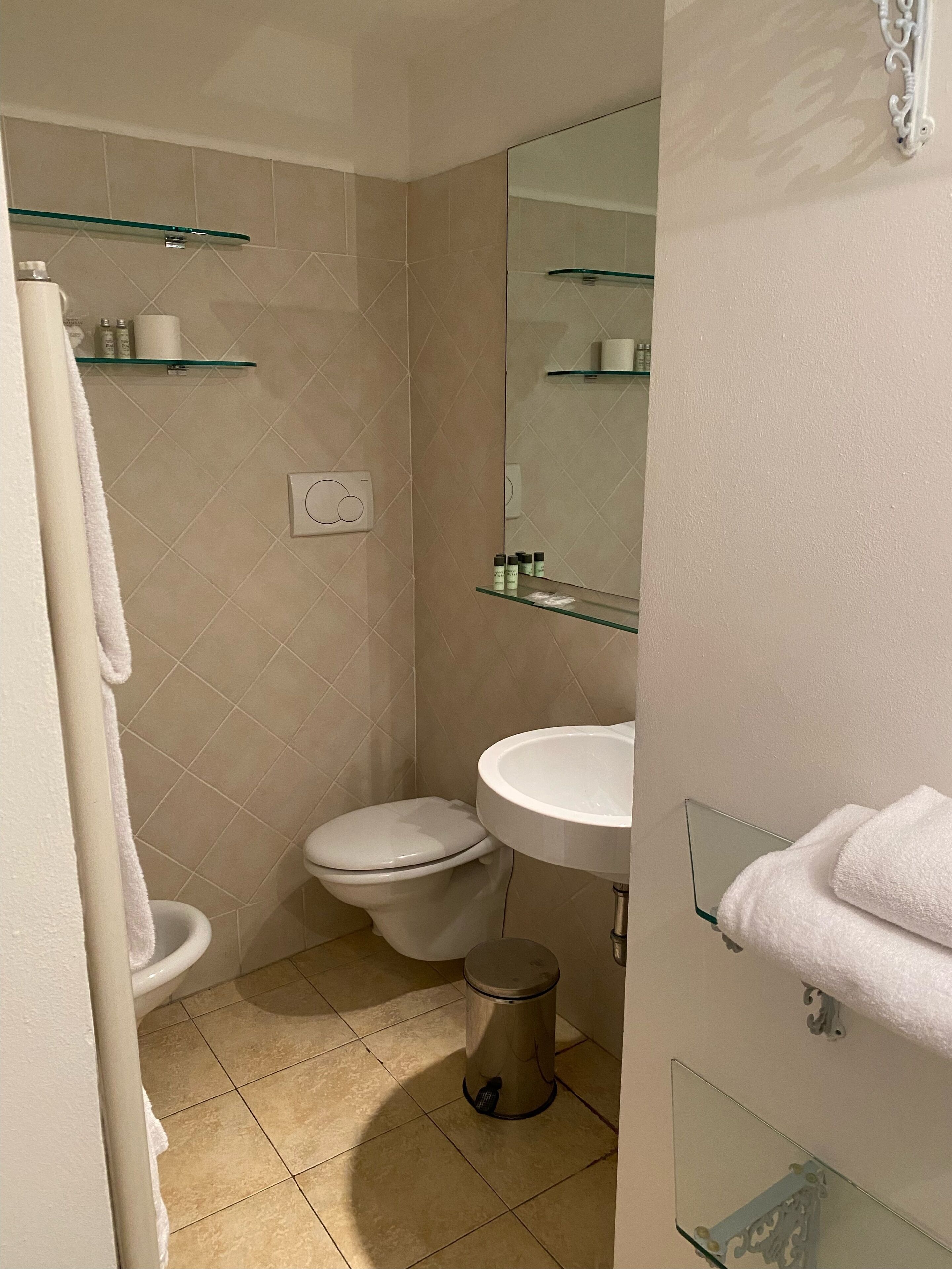 Un baño con inodoro, lavamanos y cubo de basura. Hay repisas con artículos de aseo y un espejo sobre el lavamanos.