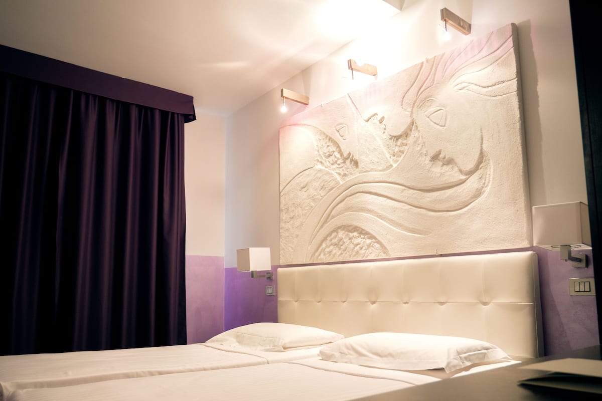 Een hotelkamer met een bed, een hoofdeinde met een structuur en een wandlamp, plus een gordijn.