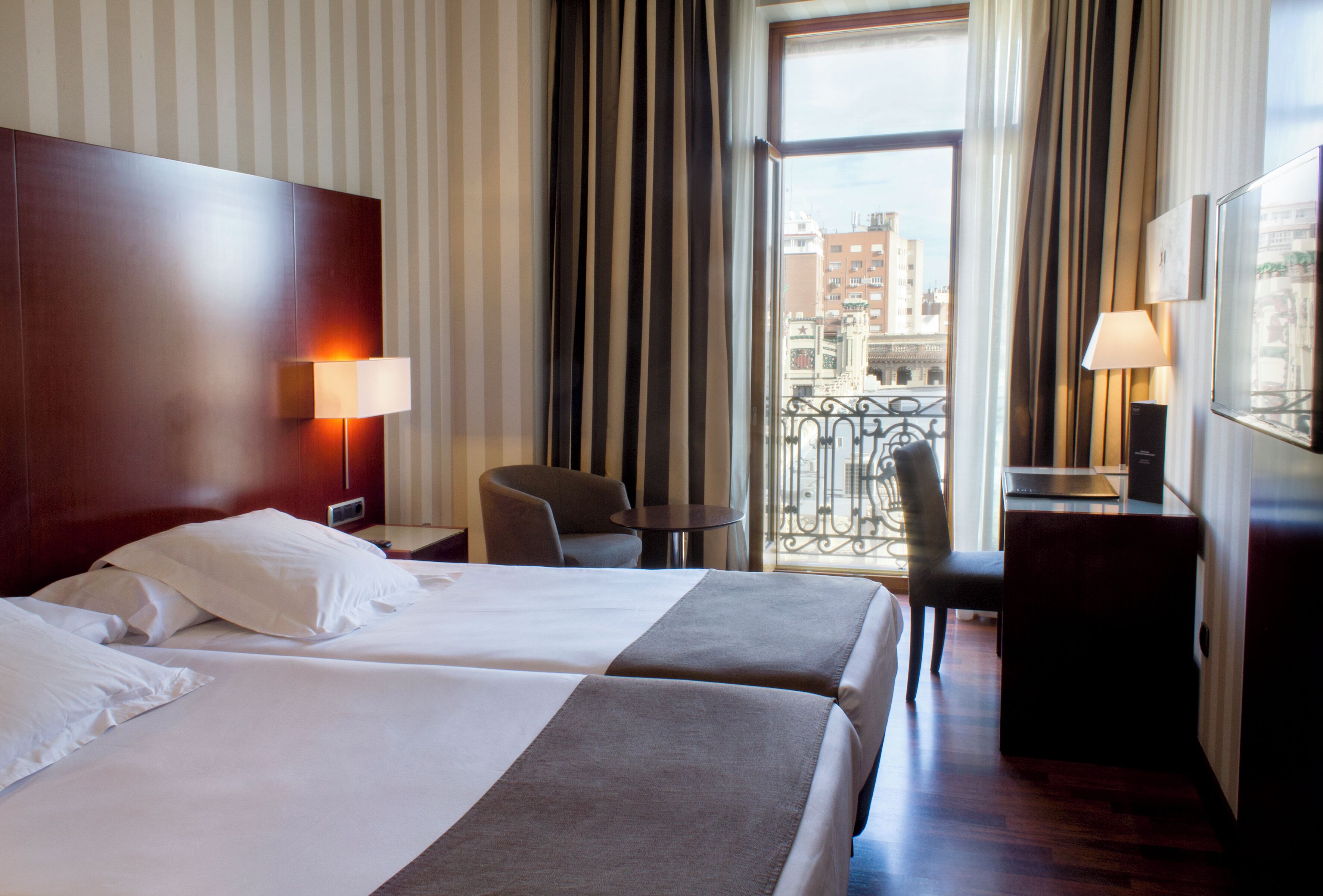 A hotel room with two beds, a desk, a chair, a small table, and a view of the city.