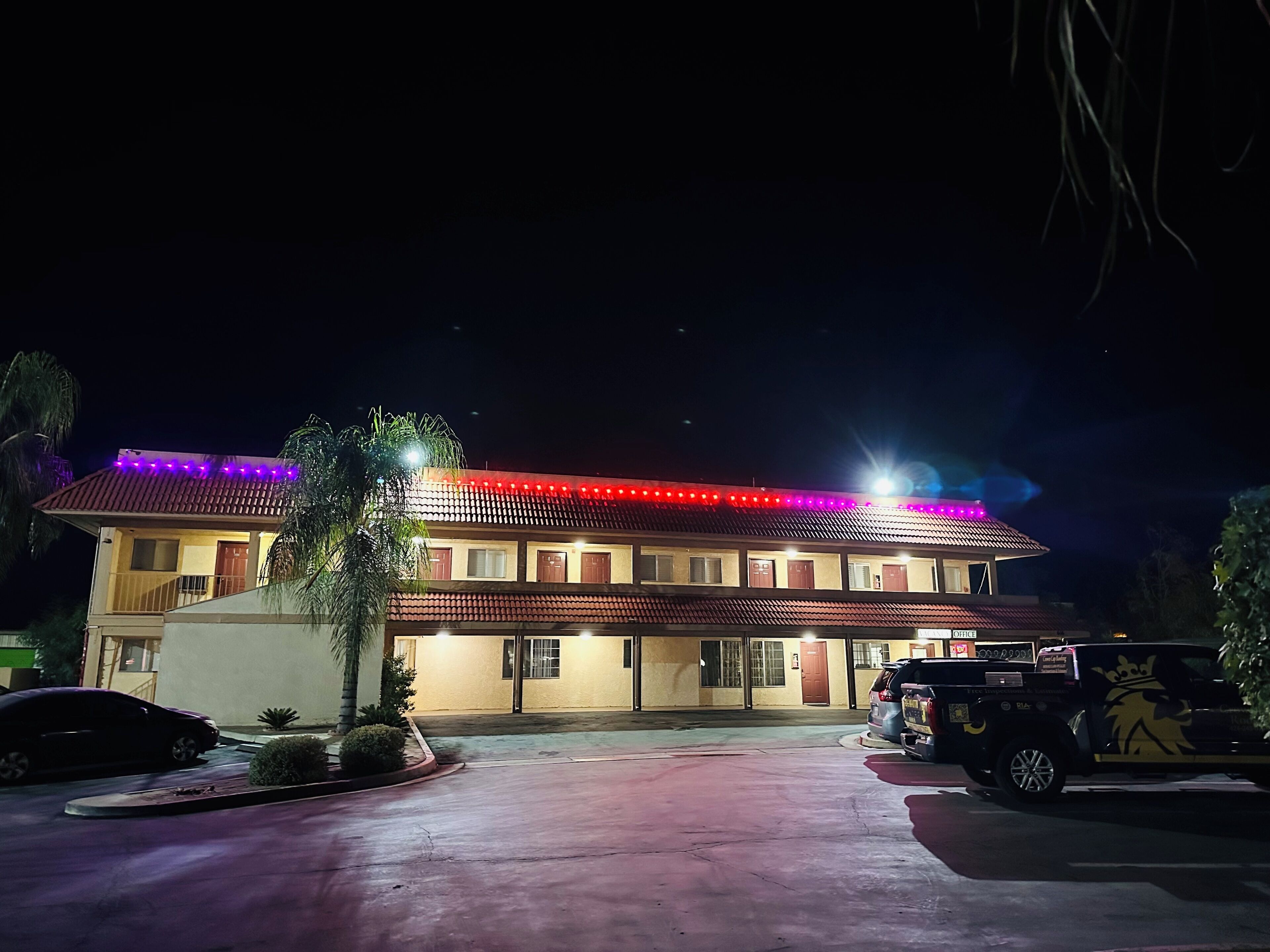 A two-story building with a red-tiled roof, multiple windows, and a parking area with a few cars.
