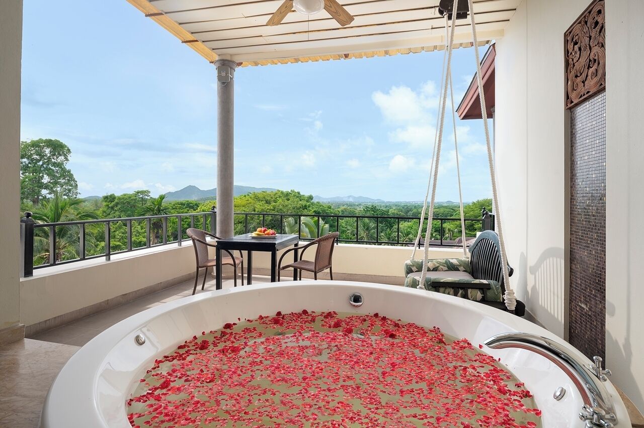 A balcony with a hot tub filled with rose petals, a table with fruit, and a view of greenery and mountains.