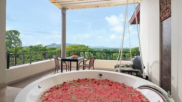 A balcony with a hot tub filled with rose petals, a table with fruit, and a view of greenery and mountains.