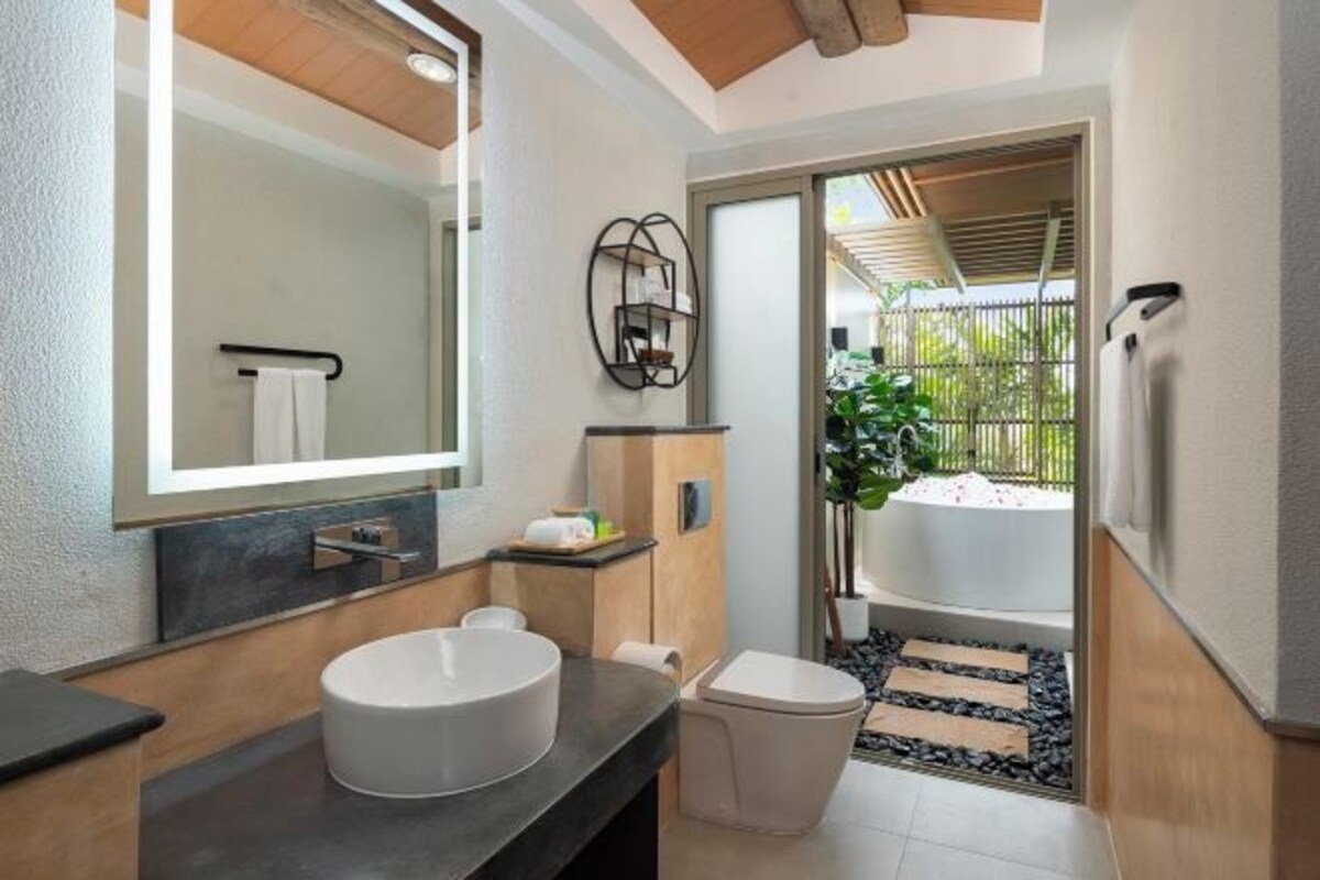 A modern bathroom with a large mirror, a round sink, and a toilet. There is a small shelf with toiletries and a towel rack above the sink.