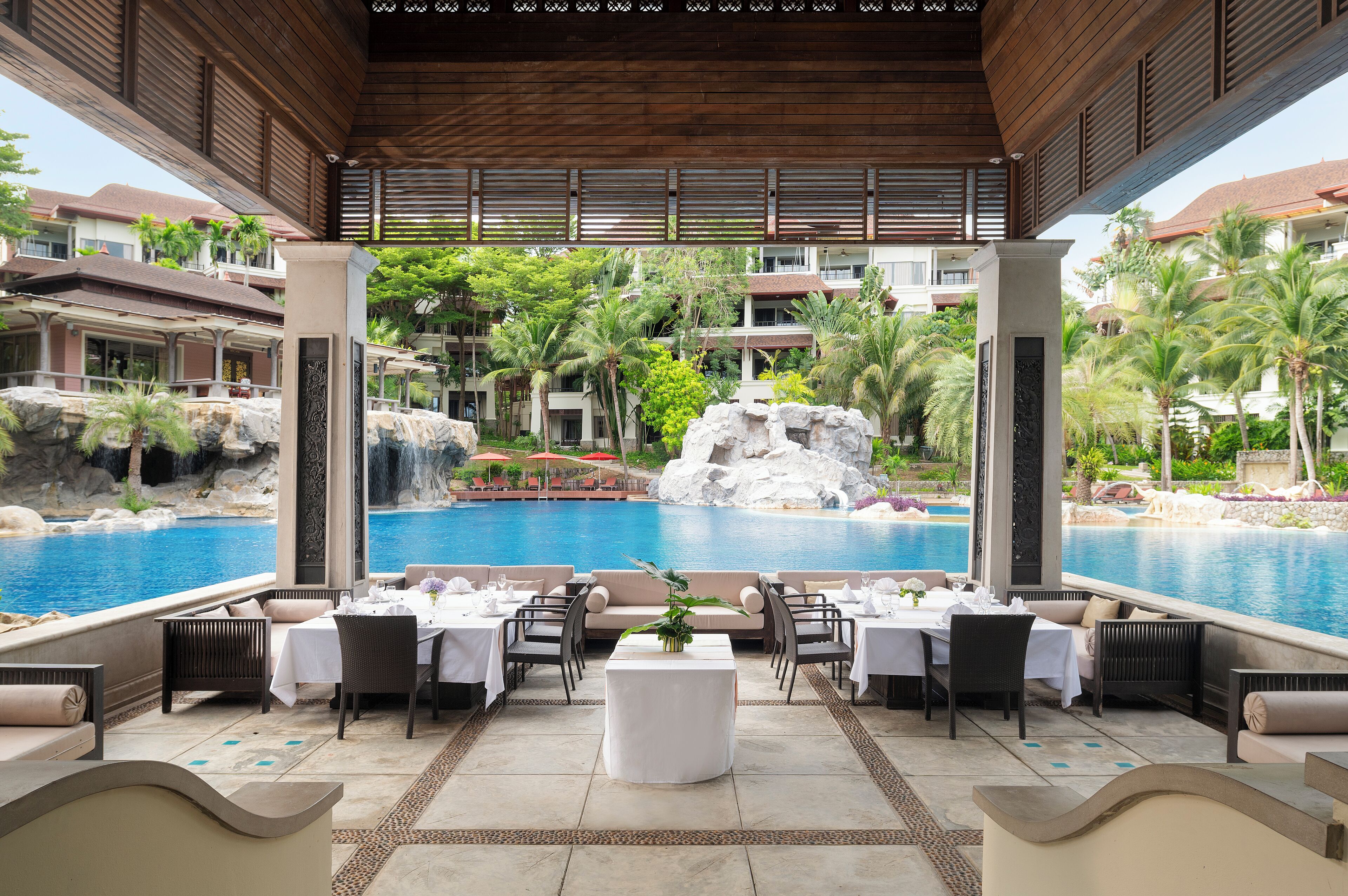 A poolside dining area with tables set for two, overlooking a large pool and a waterfall.