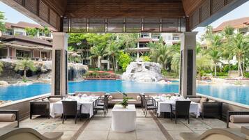 A poolside dining area with tables set for two, overlooking a large pool and a waterfall.