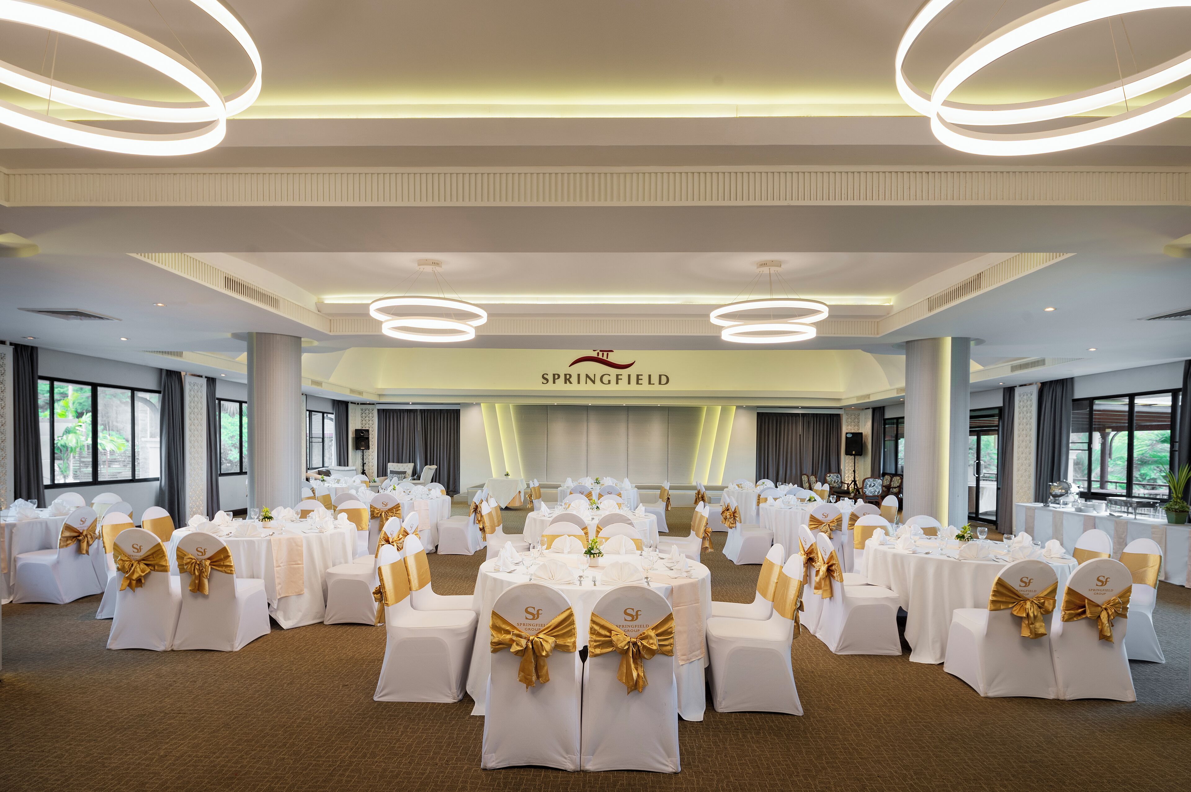 Un salón de banquetes con mesas redondas, sillas blancas y cintas doradas.