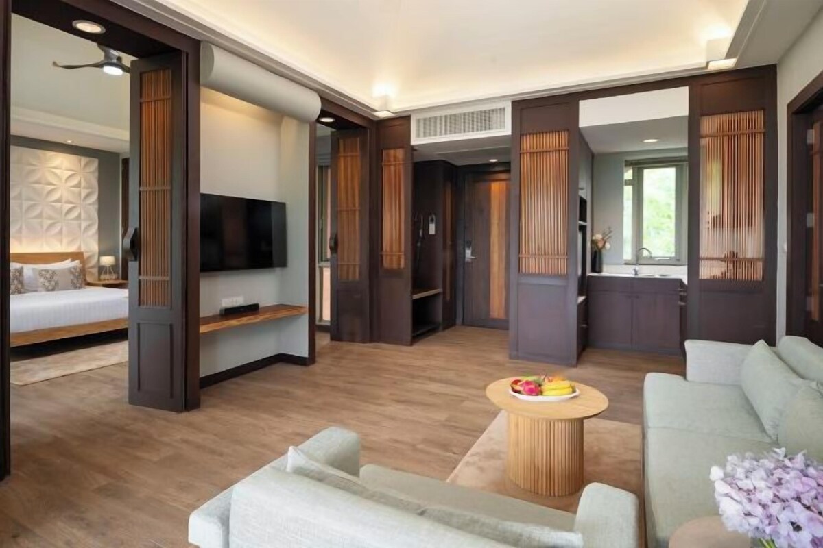 A modern living room with a sofa, a coffee table, and a TV. There is a kitchen area with cabinets and a window.
