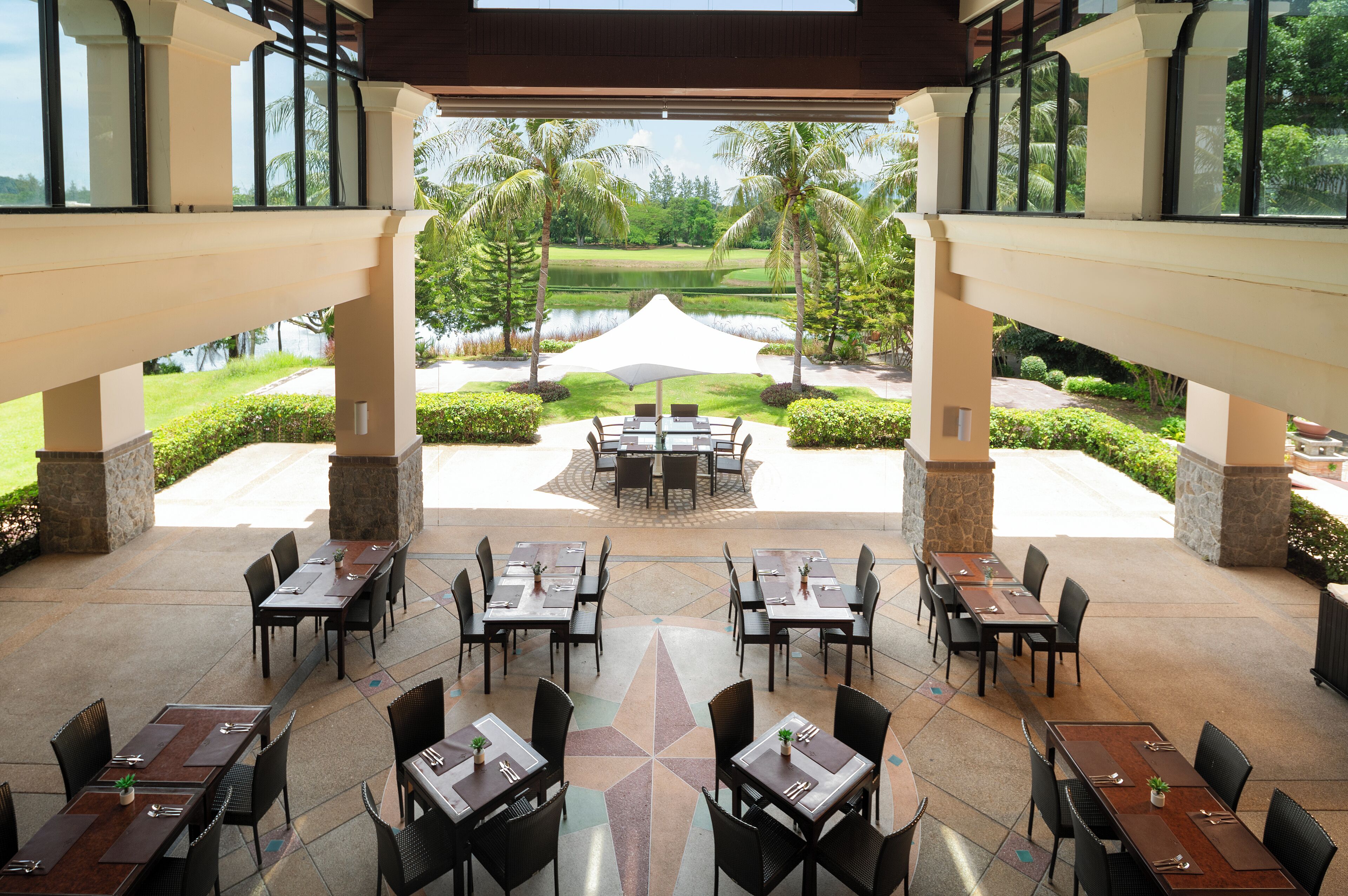 Un espacio de comedor al aire libre con mesas y sillas, vista a un campo de golf y palmeras.